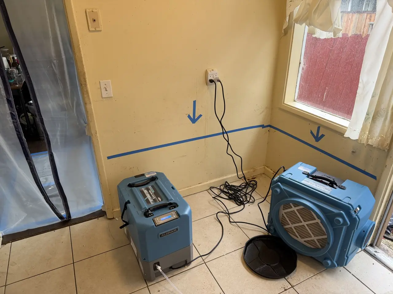Blue tape line and arrows marked along a kitchen wall, with a dehumidifier and air scrubber running