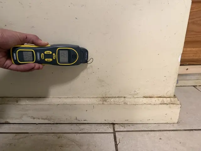 Moisture meter against the kitchen wall above the baseboard, by the brown cabinets